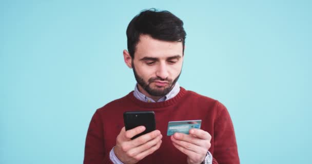 Serios homme faire une transaction en ligne à l'aide de son smartphone et une carte de crédit dans un studio avec un mur de fond bleu . — Video