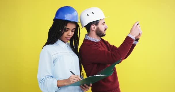 Unga afrikanska damen med dreadlocks ingenjör undertecknat dokument för nästa konstruktionsplanen med en affärsman, de båda bär en hjälm, i studion med en gul bakgrund vägg på den — Stockvideo