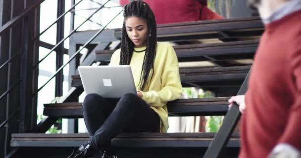 Dans l'immeuble moderne employée de bureau femme africaine assise dans les escaliers et tapant sur son carnet, à côté de son directeur de bureau marchant sur le haut des escaliers . — Video