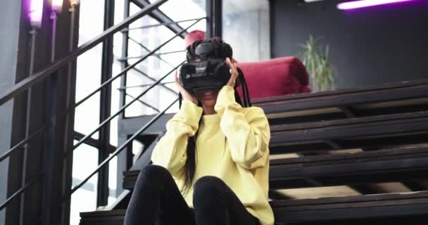 Возбужденная африканка в очках виртуальной реальности, сидящая на лестнице и играющая с большим энтузиазмом в игре, прикасаясь к программе виртуального выбора — стоковое видео