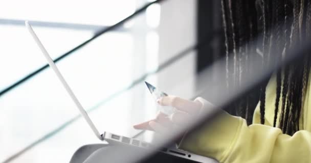 Détails close seup dame africaine avec dreadlocks à l'aide d'une carte de crédit pour faire une transaction en ligne sur son ordinateur portable, elle s'assied sur les escaliers dans un bâtiment moderne. 4k — Video