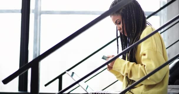 Jó kinézetű afrikai nő, ő hitelkártyával vásárolni valamit online, használja a notebook hosszú raszta ő ül a modern lépcsők az irodaház — Stock videók