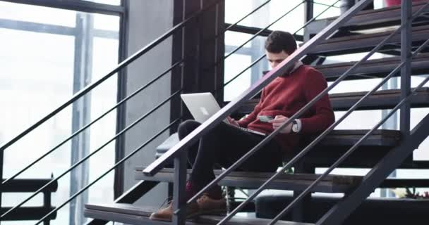 Очень счастливый и улыбчивый молодой человек офисный менеджер посреди офисного зала, сидя на лестнице и используя карточку, совершает транзакцию онлайн из ноутбука — стоковое видео
