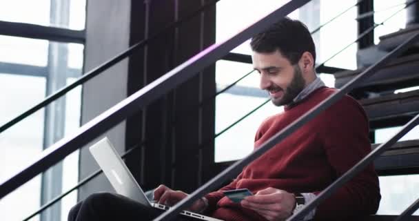 Retrato de um grande empresário sorridente sentado nas escadas do escritório e fazer um pedido on-line de notebook usando seu cartão de crédito — Vídeo de Stock