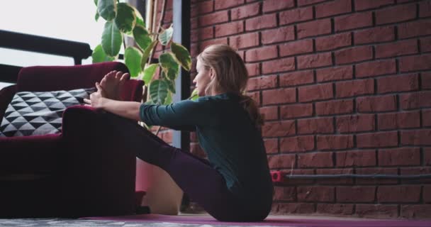 En una casa moderna en un espacio relajado señora que practica yoga por la mañana que estira las piernas y el cuerpo se siente bien usando una ropa deportiva cómoda — Vídeo de stock