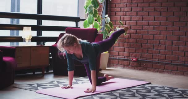 In einer urbanen Design-Dame auf einer rosa Matte, die zu Hause Yoga praktiziert, dehnt sie morgens ihren Körper, um mehr Energie für den ganzen Tag zu bekommen — Stockvideo