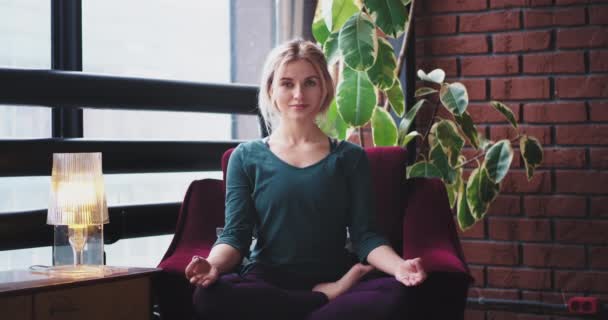 Dame pratiquant le yoga à la maison, elle est assise sur le canapé rouge et fait une posture de méditation, le matin à la maison. 4k — Video