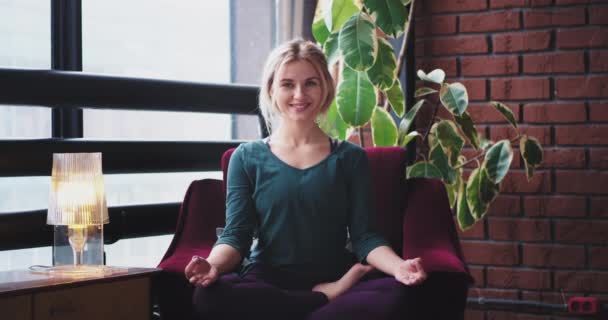 Sonriendo en un buen humor señora practicando la meditación plantea de yoga en casa en el sofá en un ambiente de sala de estar moderno. 4k. mociones lentas — Vídeo de stock