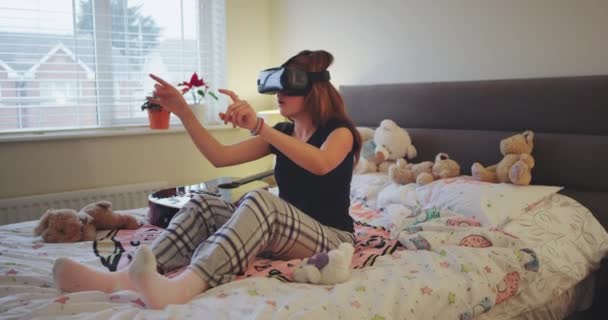 Adolescente joven carismática usando una VR para jugar algún juego virtual, ella está en su habitación usando un pijama y explorando el mundo virtual tocando virtual con el dedo el juego del programa . — Vídeo de stock