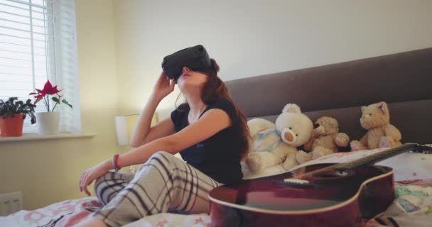 Joven adolescente jugando con unas gafas de realidad virtual que explora el mundo virtual viajando muy impresionada, en su dormitorio en la cama que lleva un pijama . — Vídeo de stock