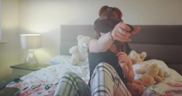 Glad och karismatisk flicka i en pyjamas på sängen spela entusiastiska med en virtual reality-glasögon hon är mycket koncentrerad och har ett uttrycksfullt ansikte. 4k — Stockvideo