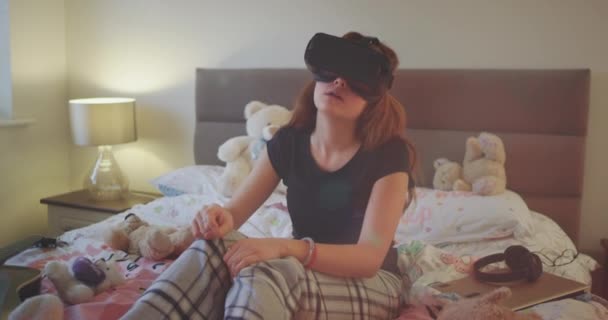 Em uma adolescente deitada na cama e usando uma nova tecnologia VR para jogar um jogo virtual, ela está muito concentrada e curiosamente . — Vídeo de Stock