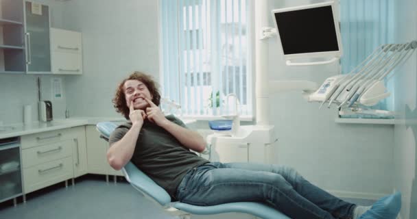 Carismático joven paciente en la clínica dental esperando a su dentista y mostrando delante de la cámara sus dientes blancos perfectos tiene un gran estado de ánimo y caras felices. 4k — Vídeo de stock