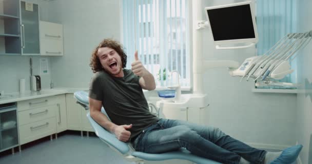 I en tandklinik rum en man patienten väntar hans läkare som sitter på tandläkare stolen och glad visar kameran en stor ut, han har ett gott humör och stort leende. 4k — Stockvideo