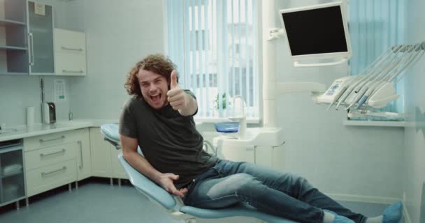 Souriant grand jeune homme dans la chambre du dentiste en attente de son examen oral, avoir une humeur parfaite montrant un grand comme devant la caméra. 4k — Video