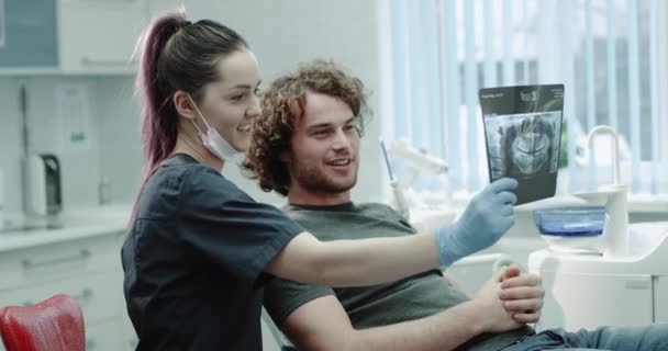 In a light dental room young dentist woman explain the result of teeth X-ray to the patient they have a friendly conversation, patient are happy for result he is sitting on dentis chair. — Stock Video