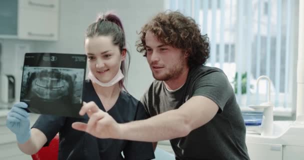 Souriant jeune homme charismatique ont un rendez-vous chez son dentiste pour obtenir ses résultats de radiographie dentaire, dentiste expliquer son problème ils sont très heureux looo à travers la radiographie. 4k — Video