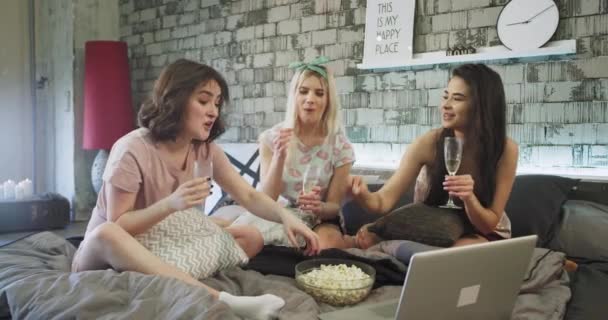 Gelukkig en charismatische multi etnische dames thuis op een modern bed van de wegens tijdgebrek grappige, drinken champagne en sommige popcorn eten tijdens het kijken naar de film op de notebook. — Stockvideo