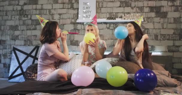 Belles jeunes filles multi ethniques soufflent les ballons tout en célébrant la fête d'anniversaire à la maison sur une chambre moderne portant un pyjama confortable . — Video