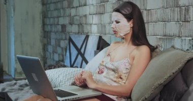 Uyku gitmeden önce pijama genç Bayan iyi görünümlü o yüzler üzerinde bir levha maskesi kullanarak bir güzellik rutin var ve maske çıkarmak için beklerken Notebook üzerinde çalışıyor