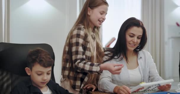 Madre muy carismática con sus dos hijos pasando tiempo juntos mientras hacen la tarea sonriendo tienen una relación amistosa, abuela de fondo leyendo un libro sentado en el sofá — Vídeo de stock