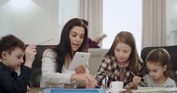 Madre madura muy expresiva mientras hace los deberes con sus hijos toma una tarjeta de crédito y comienza a pedir algo de la tableta, en una casa moderna, abuela leyendo un libro sobre el — Vídeos de Stock