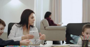 Pretty görünümlü Olgun Anne, oturma odasında modern bir evde, Büyükanne arka planda bir kitap okuma bir gadget kullanırken nasıl ödevini yapmak için ona çocuklara açıklamak