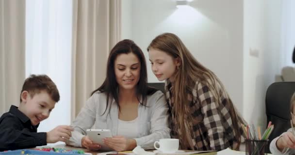 Krásná žena s bílým úsměvem dospívající matka tráví příjemný přátelský čas se svými dětmi, zatímco pracuje z domova pomocí tabletu a její děti dělají domácí úkoly