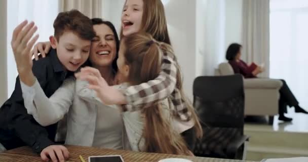 Feliz madre con sus tres hijos abrazándose juntos como un equipo amigable en la mesa de la sala de estar, mientras que la abuela leyendo un libro en el sofá en el fondo . — Vídeos de Stock