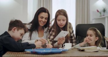 Ev ödevi zaman olgun anne beyaz ve büyük bir gülümseme ile çocuklar bazı okul projesi yapmak için yardımcı, onlar çok dostça bir takım olan bir tablet kullanarak, modern bir oturma odasında.