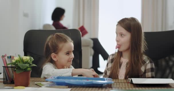 Carismática e engraçada duas irmãs brincando juntas no jogo de mesa, enquanto sua mãe passa tempo com seu filho pintando no fundo avó lendo um livro — Vídeo de Stock