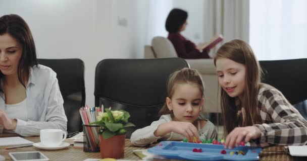 Impresionante madre madura con sus tres hijos haciendo deberes en la mesa de la cocina, pintando juntos y pasando un buen rato juntos, mientras que la abuela leyendo un libro en el fondo — Vídeos de Stock