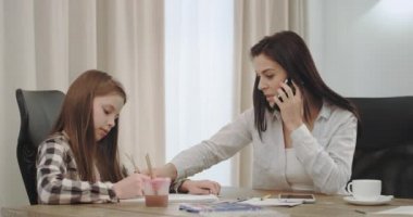 Meşgul Olgun Anne sabah evde çalışma kahve ve onu üç çocuk ile zaman harcamak bir mutfak masasında bir el sanatları zaman var, arka plan büyükanne okuma kanepede