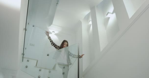 Σε ένα όμορφο μοντέρνο σπίτι πολύ φωτεινό ευτυχισμένο εννιά χρόνια κορίτσι με ένα μακρύ σγουρά μαλλιά τρέχει κάτω στις σκάλες στον πρώτο όροφο χαμογελούσε μεγάλο. 4K — Αρχείο Βίντεο