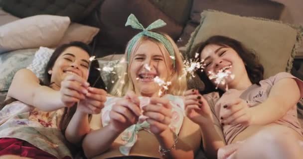 Multi etnikai hölgyek pizsama megállapításáról szóló, az ágyon egy csillogó tűzijáték előtt a kamera mosolyogva nagy őket gazdaságban, és szórakozni a Sleepover fél — Stock videók