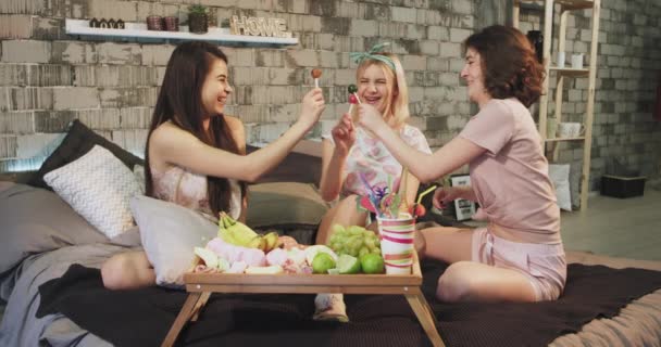 Charismatické velké usmívající se dámy si hrají s lízátka před kamerou v ložnici, která leží na posteli v pyžamu. — Stock video