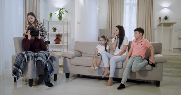 Бабушка с внуками и зрелая мать проводят время вместе на диване, исследуя новые очки виртуальной реальности, которые племянница надела на свою бабушку виртуальную машину. . — стоковое видео