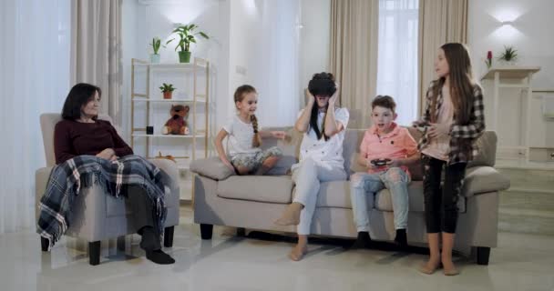 A big family mother with three kids and granny have a good time together they playing on video game, mother using a VR to exploring the virtual world, granny impressed looking at them, they sitting — стоковое видео