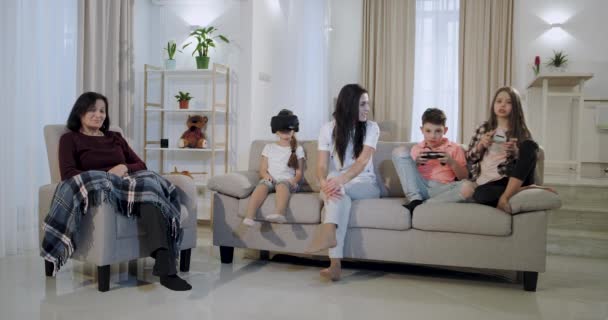 Η γιαγιά με τα εγγόνια της και μια ώριμη μητέρα που περνούν το χρόνο τους μαζί στο σαλόνι τα παιδιά παίζουν σε ένα βιντεοπαιχνίδι και χρησιμοποιώντας ένα VR για να εξερευνήσετε τον εικονικό κόσμο — Αρχείο Βίντεο