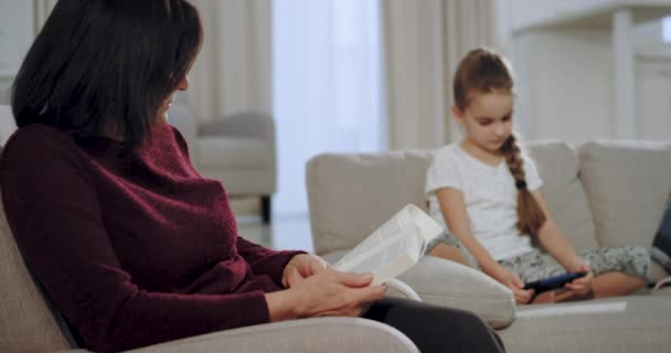 Karismatisk liten flicka som spelar på en smartphone spel medan hennes farmor koncentrerade läsa en bok på soffan i ett stort rymligt vardagsrum — Stockvideo