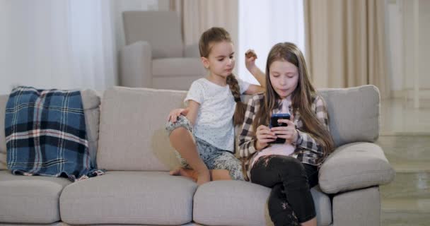 Two charismatic sisters sitting on the sofa and eating some biscuits and playing on a smartphone , spending a fun time together — Stock Video