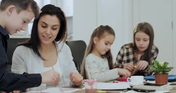 Tres niños pequeños después de la escuela haciendo la tarea en la sala de estar en una mesa grande junto con su madre madura y hermosa — Vídeos de Stock