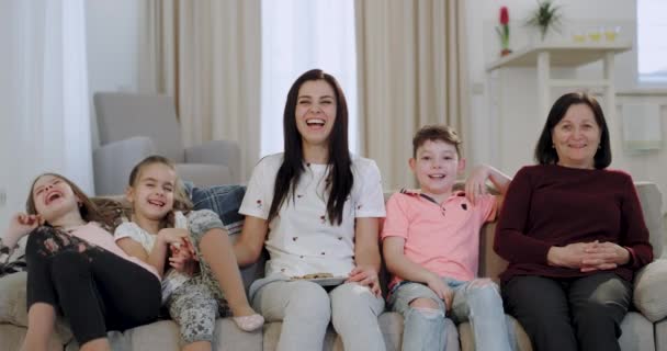 Glimlachend groot aantrekkelijk oma haar dochter en kleinkinderen voor de camere tijdens het kijken naar de TV zittend op de Bank, ze zijn blij genieten van de tijd samen — Stockvideo