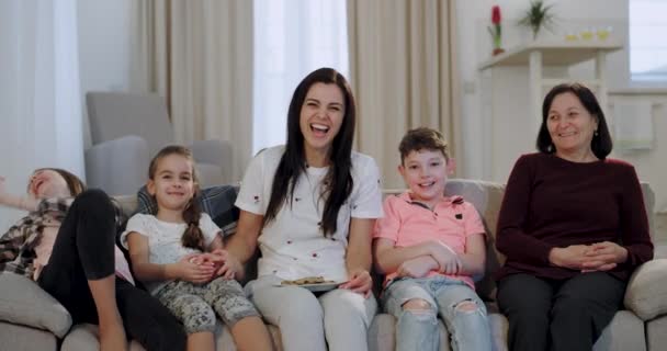 Gelukkig lachende charismatische kinderen met hun moeder en oma kijken naar een komediefilm op de TV voor de camera terwijl zittend op de Bank — Stockvideo
