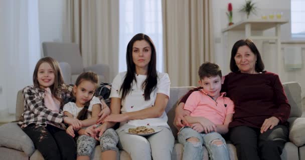 Geconcentreerde en grappige familie oma met haar mooie dochter en kleinkinderen kijken naar een komediefilm op de TV ze glimlachen groot en genieten van de tijd samen — Stockvideo