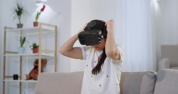 Attraente donna che indossa occhiali di realtà virtuale mentre è seduta sul divano, pronta a viaggiare in tutto il mondo con una VR — Video Stock