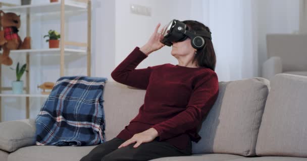 Mujer vieja concentrada usando por primera vez unas gafas de realidad virtual mientras está sentada en el sofá, está muy impresionada con este nuevo desarrollo tecnológico — Vídeo de stock