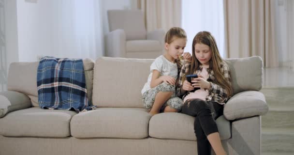 Две симпатичные сестры, сидя на диване, играют в игру на смартфоне в просторной современной гостиной, очень сосредоточенно проводя время вместе — стоковое видео