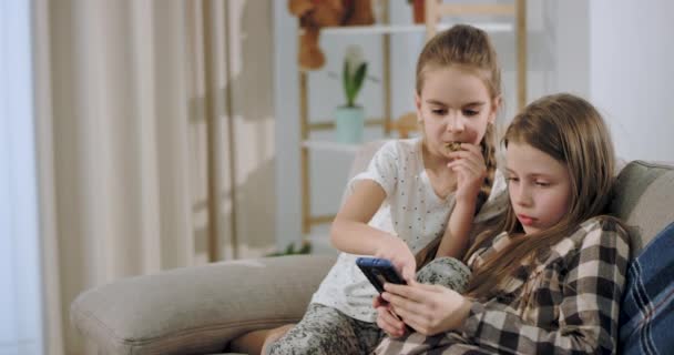 Closeup carismático e bonito duas irmãs jogando juntos em um jogo usando um smartphone enquanto sentado no sofá . — Vídeo de Stock