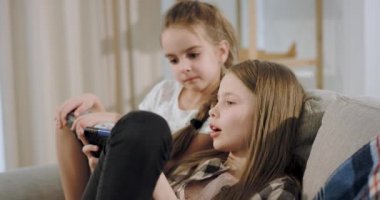 Kanepede otururken bir akıllı telefon kullanarak bir oyun üzerinde birlikte oynayan iki karizmatik kız portresi yanlarında bir kitap okuma ve torunları bakarak. 4k